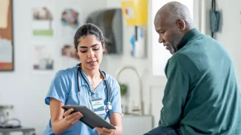 A nurse in blue scrubs with a stethoscope consults with a Black patient in a clinic, using a tablet. Warm, professional healthcare scene highlighting patient care and medical teamwork.