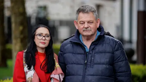 PA Media Natasha Butler and Harry Gargan. Natasha has long dark hair, wearing glasses, a red coat and red top and pattered scarf. Harry has short grey hair and moustache, wearing a navy puffer coat and light blue top. 