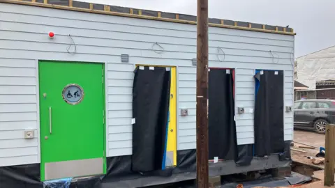 A container shaped toilet block under construction. It has a green door for disabled and family access , and several other coloured doors for regular users.