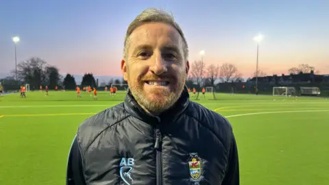 Aidy Blake smiles at the camera. He has short grey hair and a short beard. He wears a navy coat with the team's logo on, and stands with a football pitch behind him with players in the distance. The sun is settling in the distance, turning the sky pink and purple. 