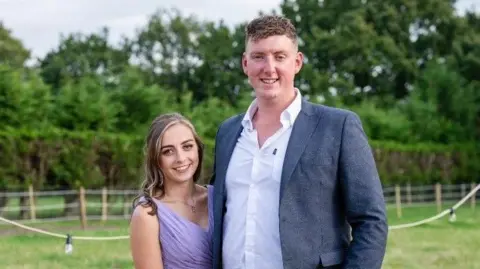 Staffordshire County Council A woman and a man are standing outdoors. The woman is wearing a purple dress and the man is wearing a suit with open collar. They are smiling at the camera.