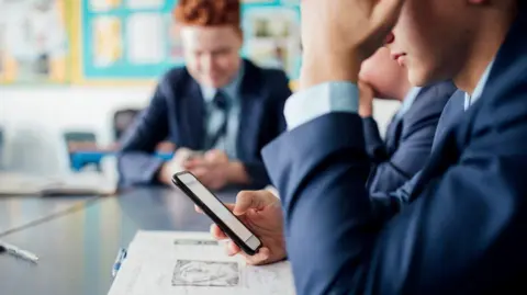 Generic picture of a boy in school uniform looking at his mobile phone in a classroom while he has schoolwork in front of him. Another boy further away is doing the same