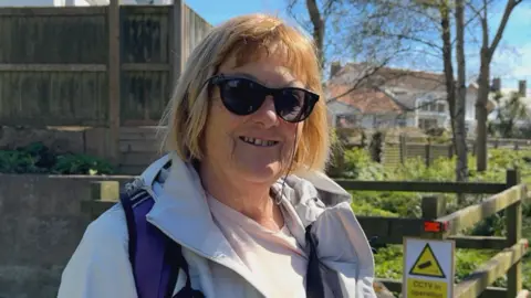 A woman wearing sunglasses and a light jacket stands outdoors near a wooden fence and greenery, with houses and trees visible behind her. A small sign on the fence reads ‘CCTV in operation