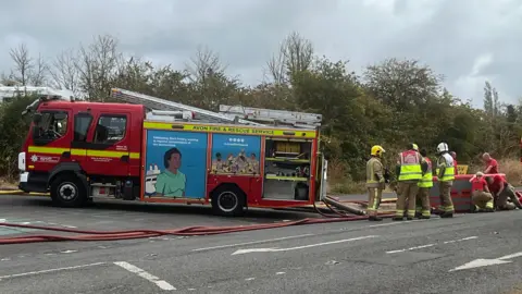 Two fire engines are parked on a road as firefighters in uniform stand next to them.
