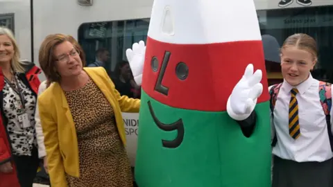 Eluned Morgan, in a yellow jacket and black and white top, next to Mistar Urdd, a white, red and green mascot