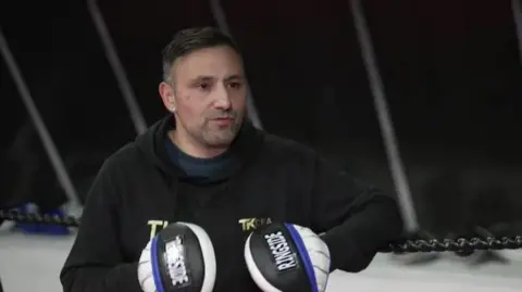 A boxer stands against some ring ropes wearing boxing gloves. He is wearing a black hooded top and looking to the side of the camera.
