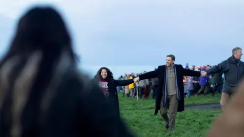 Reuters People smiling and joining hands in a circle around a field. A crowd can be seen behind the circle.
