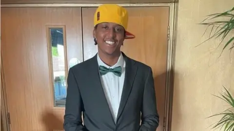 Family handout Eddie Kinuthia is standing in front of brown wooden swing doors. He is wearing a yellow baseball cap, a grey suit and green bow tie 