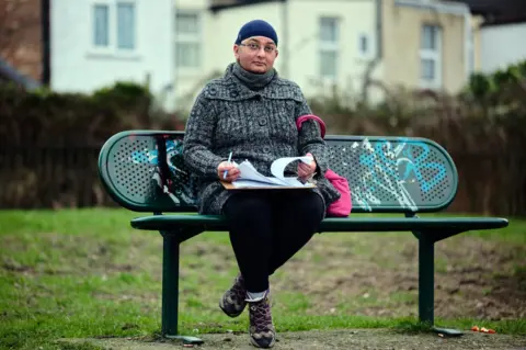 Kiran sits on a bench in a park outside the library. She is holding a petition she organised to try and help save the library from closure