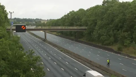 The northbound and southbound carriageway of the M1 motorway with no traffic. A man is stood on one side of the road wearing a fluorescent yellow jacket.