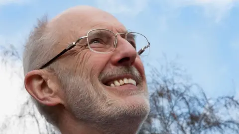 Paul Andersen A close-up of Paul Andersen, who has short grey hair and beard and wears silver-rimmed glasses. He is smiling and looking up at a blue sky.