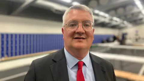 Elizabeth Baines/BBC A man with white hair wears a black suit and red tie. Behind him a sports hall is blurred.