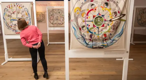 Alan Richardson A woman with short hair, a pink top and black trousers looks at the panel of the Dundee Tapestry inside a museum. Other panels from the tapestry are visible in the room.