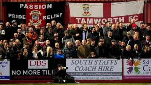 Tamworth Football Club Tamworth fans in the stands. There are sponsorship boards around them and flags in the background.