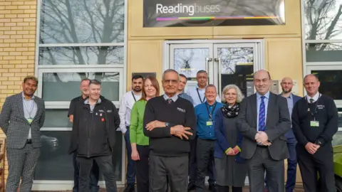 Reading Buses Mr Singh stands outside the Reading Buses office building surrounded by his colleagues. The group are all looking at the camera, with Mr Singh standing in the centre wearing his uniform - a blue collared shirt, tie and black jumper with grey formal trousers. There is a beige brick building behind the group with a Reading buses sign on it.