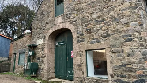 BBC Exterior of Sark Electricity's HQ. It is a stone building with a couple of windows and a double wooden green door. There is also an old green petrol pump by the wall.