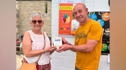 Sharon Scrivens A woman wearing a white vest top, presenting a bottle of sun cream to a man on the right wearing a yellow top. Both are smiling and looking at the camera.