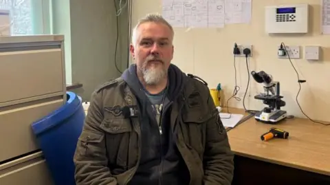 A man with grey hair and a beard sits in an office. He is wearing a green jacket and a blue hooded jumper. A desk is visible behind him with a microscope and paperwork on the surface.