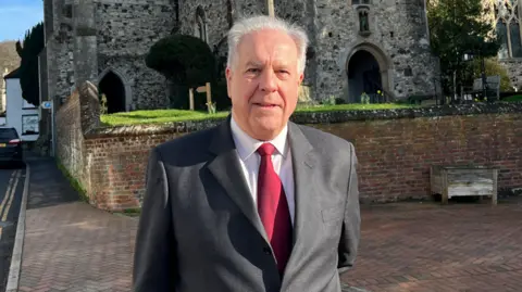 BBC/Phil Harrison A man wearing a suit. He is stood in front of a church. 
