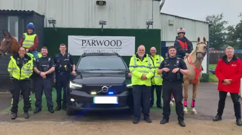 Surrey police officers stand around a car, with two riders and a sign reading "Parwood Equestrian Centre"