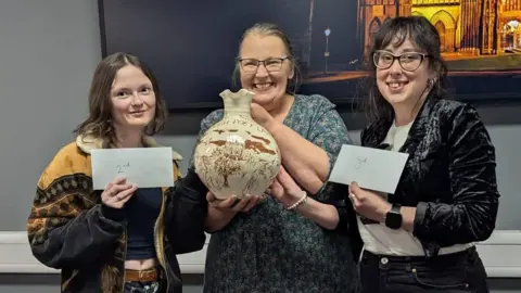 Emily Steele/Toby Wood Three finalists, with Hilary Steele in the middle, smile and hold their prizes. Hilary is holding a jug. The second and third-placed poets, both women, are holding envelopes.