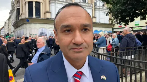 A man with short black hair, wearing a light blue shirt, a red tie and a navy suit. There is a crowd of people behind him. 