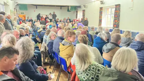 Dozens of people are sitting in a community hall in rows. 
