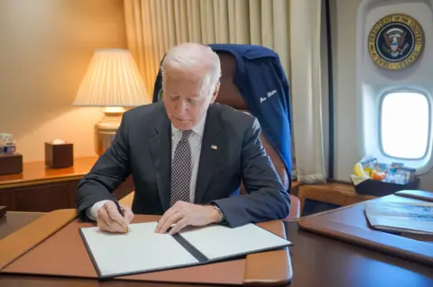 X Presiden Joe Biden duduk di mejanya di Air Force One yang menandatangani pengampunan. Dia mengenakan lencana bendera AS di kerahnya. Ada jaket biru di belakang kursinya dengan namanya ditulis di depan. Segel Presiden Amerika Serikat berada di atas jendela pesawat di belakangnya. 