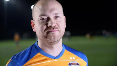 Jamie Niblock/BBC Freddie Kizis-Stevens is wearing a yellow and blue football shirt, with a Sands United crest. He is standing on a football pitch.