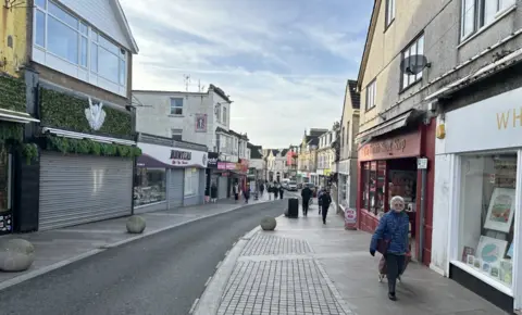 A town street with shops on eitehr side and a woman with a shopping trolley on the right with other people in the background.