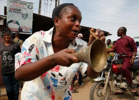 EPA Woman ringing a bell in Lagos at a polling unit. There are other people behind her on the road.