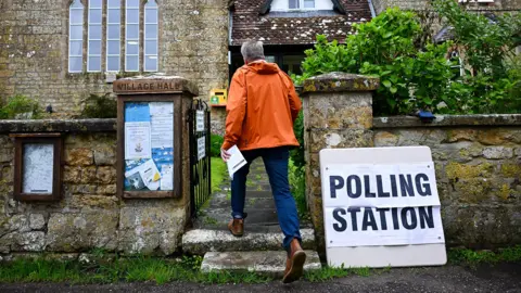 A man walking into a polling station in a rural church . He is seen from behind walking through a gate and wearing an orange coat 