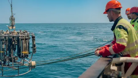 Three men in high vis jackets, wearing hard hats are pulling in the instruments, that measure the AMOC, from the ocean using ropes.