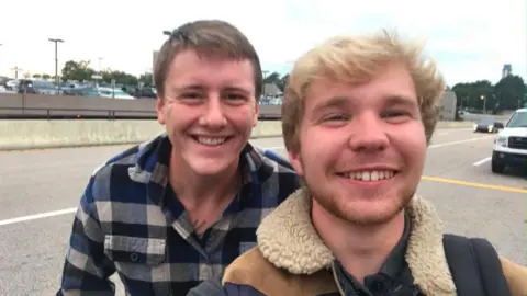 Ash Carter Tristan Torry is on the left standing slightly behind Ash Carter. Tristan has short brown hair and is smiling, and wearing a blue checked flannel shirt. Ash has blonde hair, also smiling, and is wearing a sheepskin style coat. There is a road behind them, and a car park beyond that. 