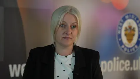 The image shows a woman with chin length blonde hair seated indoors in a formal interview-style setting, wearing a dark blazer over a light, patterned top. The person is positioned in the foreground, facing the camera, with a softly blurred background. Behind them is a branded backdrop featuring the West Midlands Police crest and partial website text