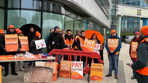 Resident Doctors go on strike and are standing outside a Queen Elizabeth Hospital and Birmingham in December 2025 .They hold orange signs with a BMA logo. Signs include 'doctors need jobs now' and 'patients need doctors, doctors need jobs'.