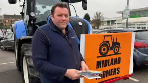 A man in a blue sweater standing in front of a tractor with an orange sign saying Midlands farmers