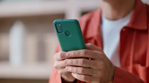 Getty Images A person wearing a bright, orange jacket and white t-shirt types on a smartphone held in their hands.