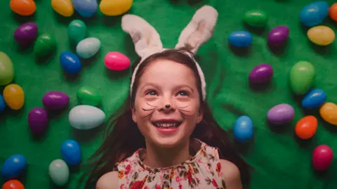 A young girl lies down on a green background with multi-coloured eggs dotted around her head. She has bunny ears on and whiskers painted on her face. She has a flowery dress on and long brown hair.