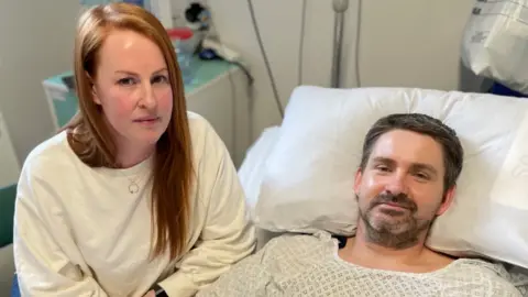 A man with short brown hair and a beard wearing a hospital gown and lying on a hospital bed. Next to him is a woman with long red hair, wearing a white jumper. Both of them are looking at the camera with serious expressions.