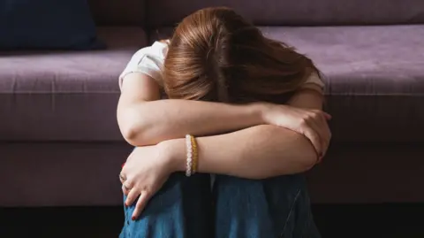 A stock image of a woman, with her head on her knees and her arms across her. A sofa is behind her, and she has on two bracelets. She has long dark hair.