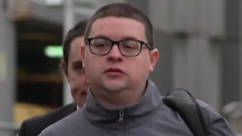 BBC Ieuan Crump is wearing a grey turtleneck jacket, black glasses and a black bag strap over his shoulder. 