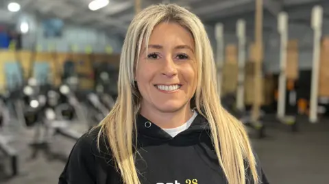 Natasha Daryaie has long blonde hair and is standing in a gym. She is wearing a black hoodie. 