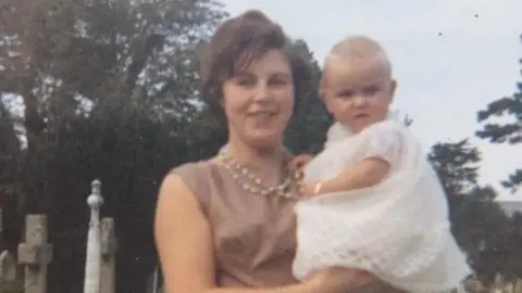 Family photo An old photo of Sandie Bowen as a young mum, holding her baby daughter Anita in her arms. Sandie is wearing a pink dress and colourful bead necklace and has short brown hair. Baby Anita is in a white Christening dress.