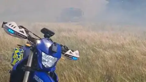 A blue, yellow and white motorbike parked up in an dry straw field with smoke in the background.