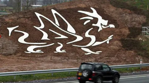 The eastbound side of the A494 dual-carriageway in Flintshire showing work being completed on a white dragon shape cut into a sloping embankment. The design is filled with white stone so that it stands out to passing motorists. A digger can be seen on the muddy ground.
