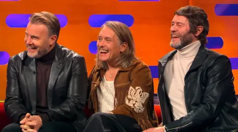 Take That/Netflix Gary Barlow, Howard Donald and Mark Owen are laughing as they sit on a couch during a TV interview for the The Graham Norton Show.