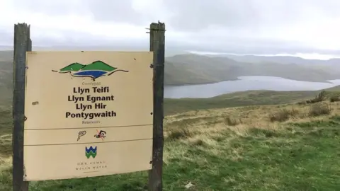 A sign for the Teifi Pool reservoir with the body of water see in the background surrounded by grassy fields.