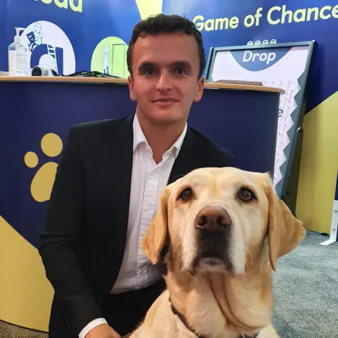 Submitted A man with short curly dark hair is wearing a dark suit and white, open-necked shirt. He is crouching with a golden Labrador dog in front of a blue and yellow charity stall. 
