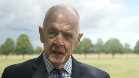 Jamie Niblock/BBC A bald man stands outside on a sunny and partly cloudy day. He is looking away from the camera as he talks. He wears a dark blue suit with a white and blue checked shirt and a purple and blue tie. 